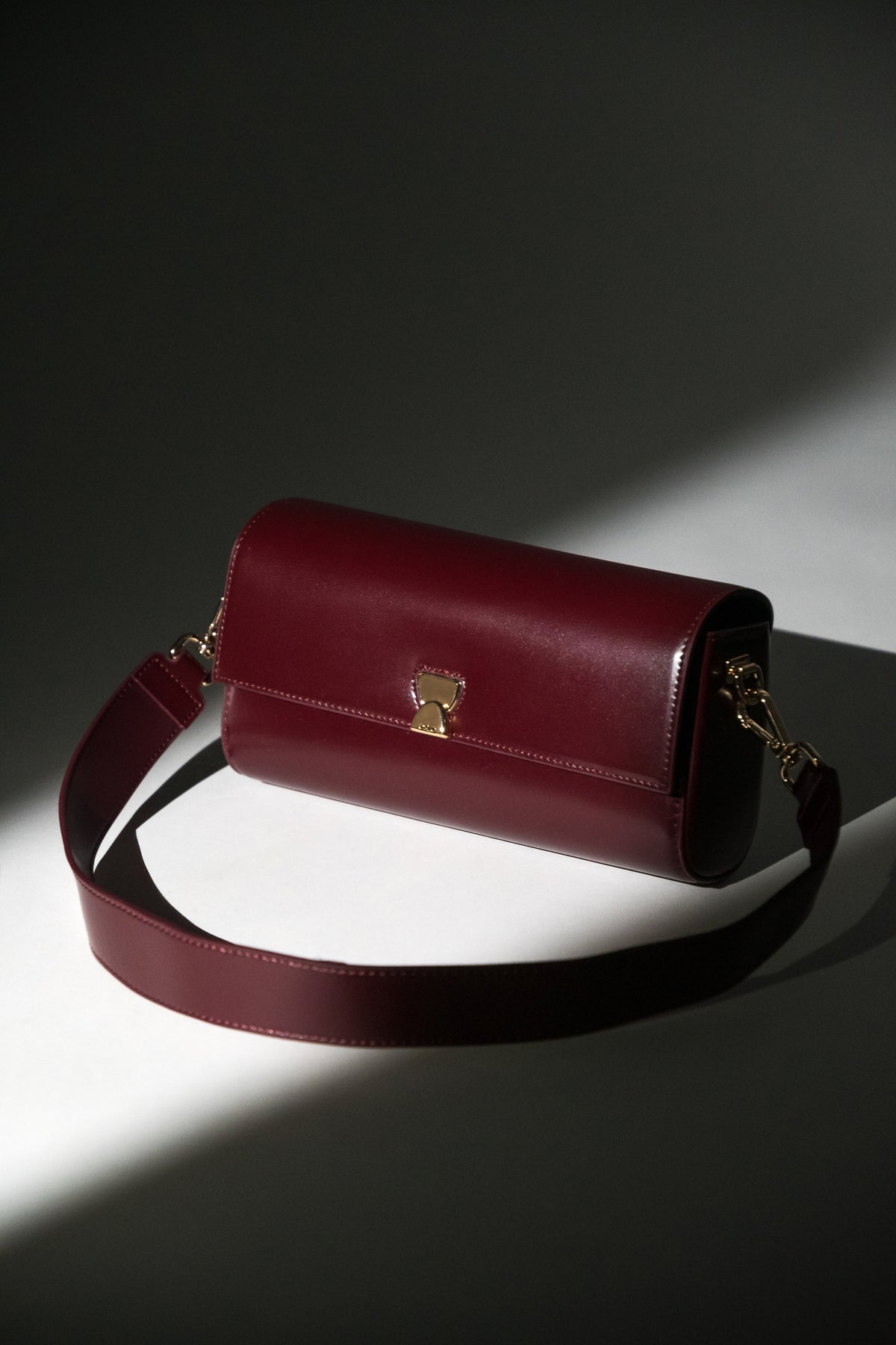 Künstlerische Aufnahme der bordeauxroten Leder-Clutch mit breitem Schulterriemen im Spotlight
