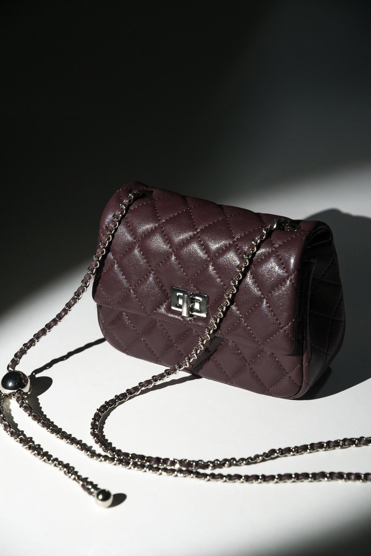 Künstlerische Studioaufnahme der bordeauxroten Damen-Stepptasche mit silberner Kette im dramatischen Licht