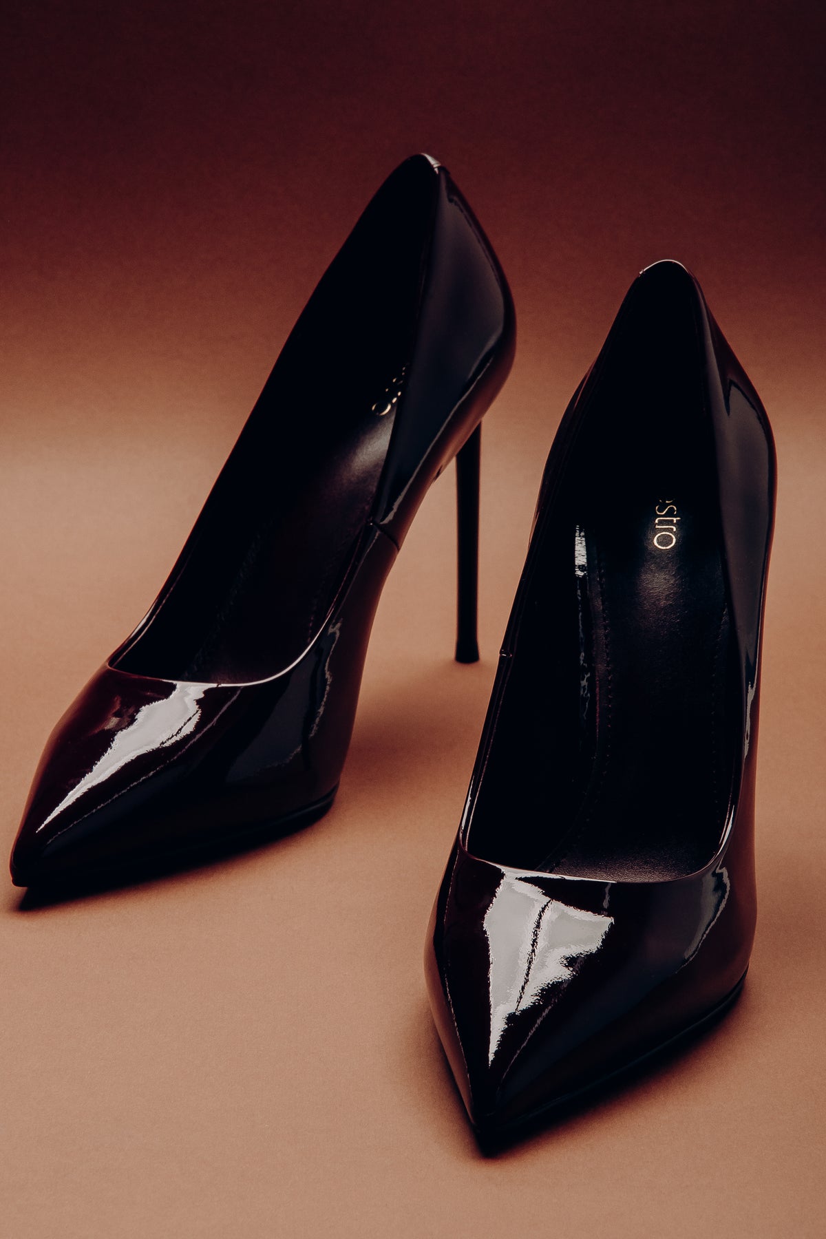 Stimmungsvolle Aufnahme zweier schwarzer Damen-Stilettos aus glänzendem Lackleder vor warmem Studiohintergrund