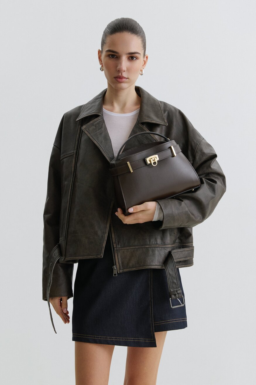 Model in dunkelbrauner Lederjacke präsentiert die dunkelbraune Handtasche in Kofferform 