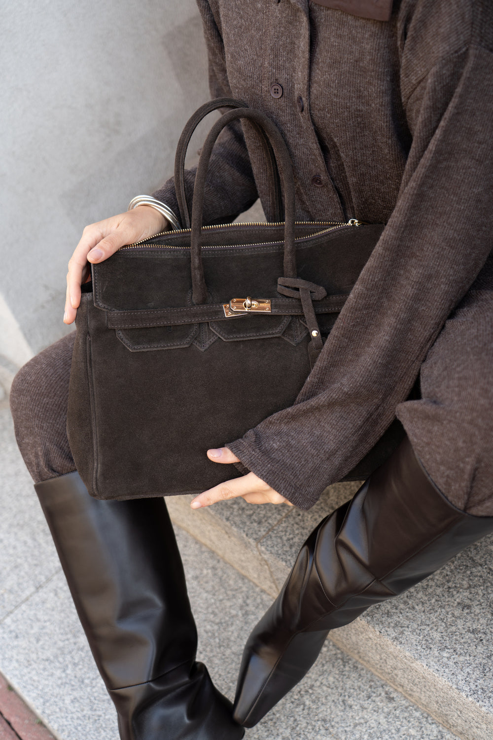 Lifestyle-Nahaufnahme: Model sitzt auf Steinstufen und hält die dunkelbraune Damen-Henkeltasche aus echtem Veloursleder auf dem Schoß