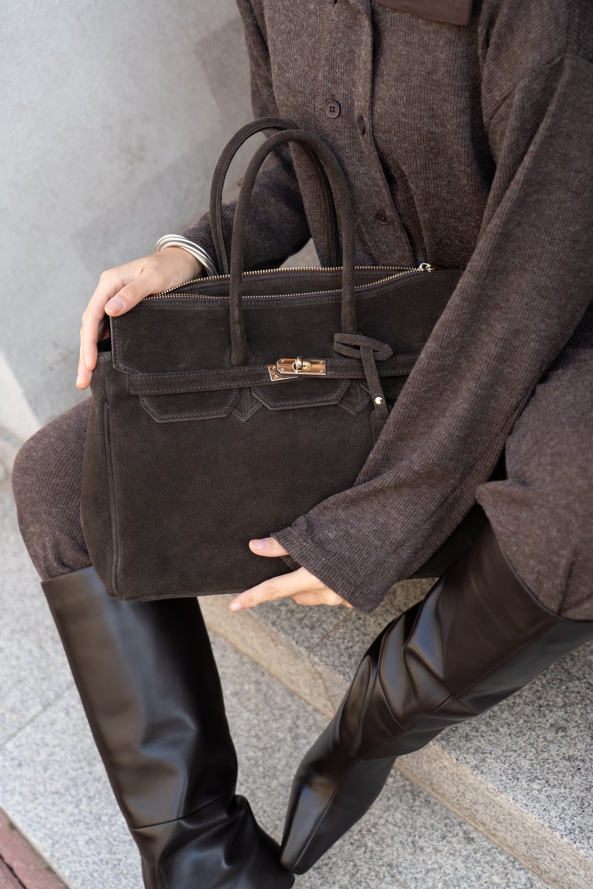 Lifestyle-Nahaufnahme: Model sitzt auf Steinstufen und hält die dunkelbraune Damen-Henkeltasche aus echtem Veloursleder auf dem Schoß
