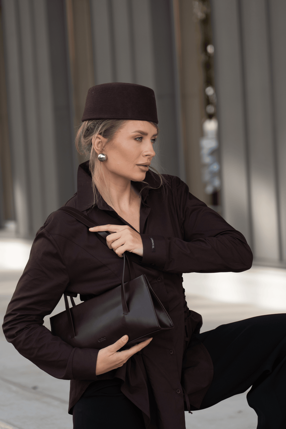 Lifestyle-Aufnahme einer Frau im dunkelbraunen Outfit mit passender Kopfbedeckung, die die dunkelbraune Leder-Schultertasche von Estro hält