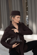 Lifestyle-Aufnahme einer Frau im dunkelbraunen Outfit mit passender Kopfbedeckung, die die dunkelbraune Leder-Schultertasche von Estro hält