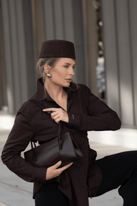 Lifestyle-Aufnahme einer Frau im dunkelbraunen Outfit mit passender Kopfbedeckung, die die dunkelbraune Leder-Schultertasche von Estro hält