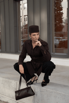 Outdoor-Lifestyle-Aufnahme einer sitzenden Frau im dunkelbraunen Outfit, die die Schultertasche auf einer Betontreppe präsentiert