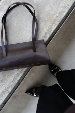 Draufsicht aus der Vogelperspektive auf die dunkelbraune Leder-Schultertasche, die auf Betontreppen neben schwarzen Hosen und Schuhen steht