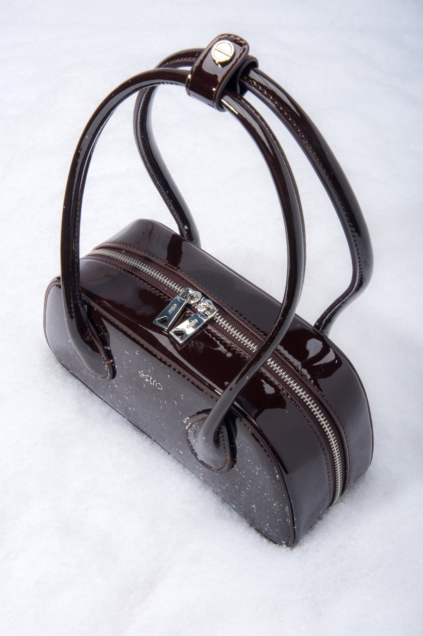 Detailaufnahme des Logo-Pins am Griff der dunkelbraunen Lackledertasche.