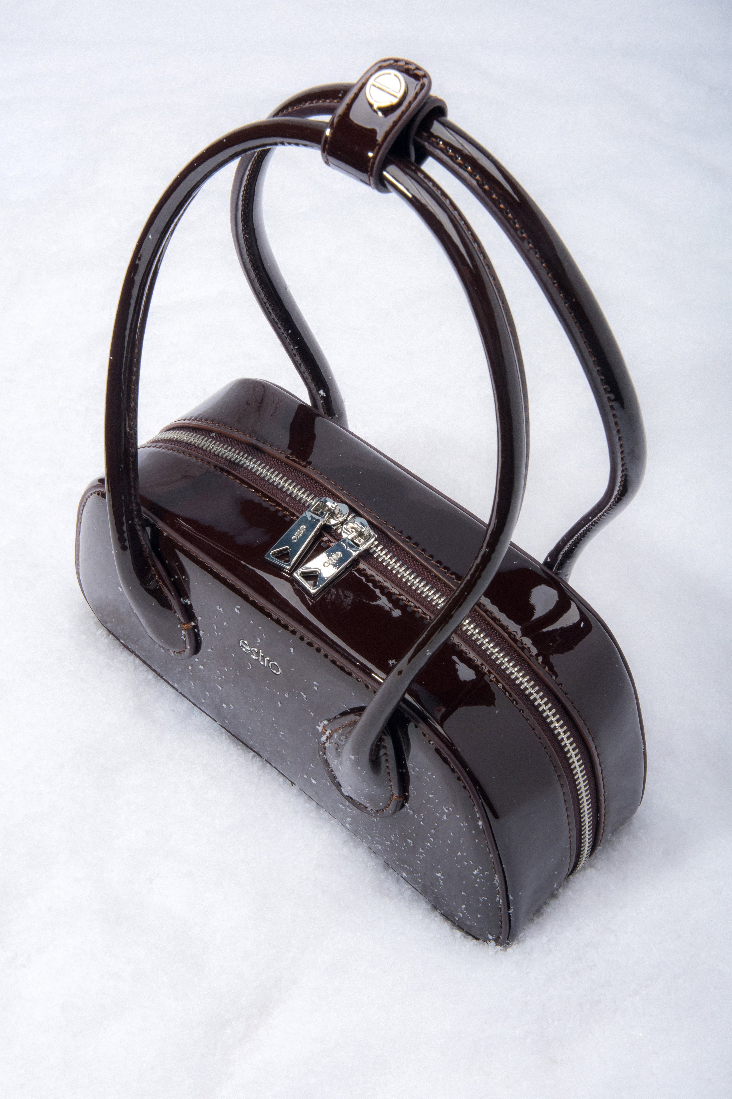 Detailaufnahme des Logo-Pins am Griff der dunkelbraunen Lackledertasche.