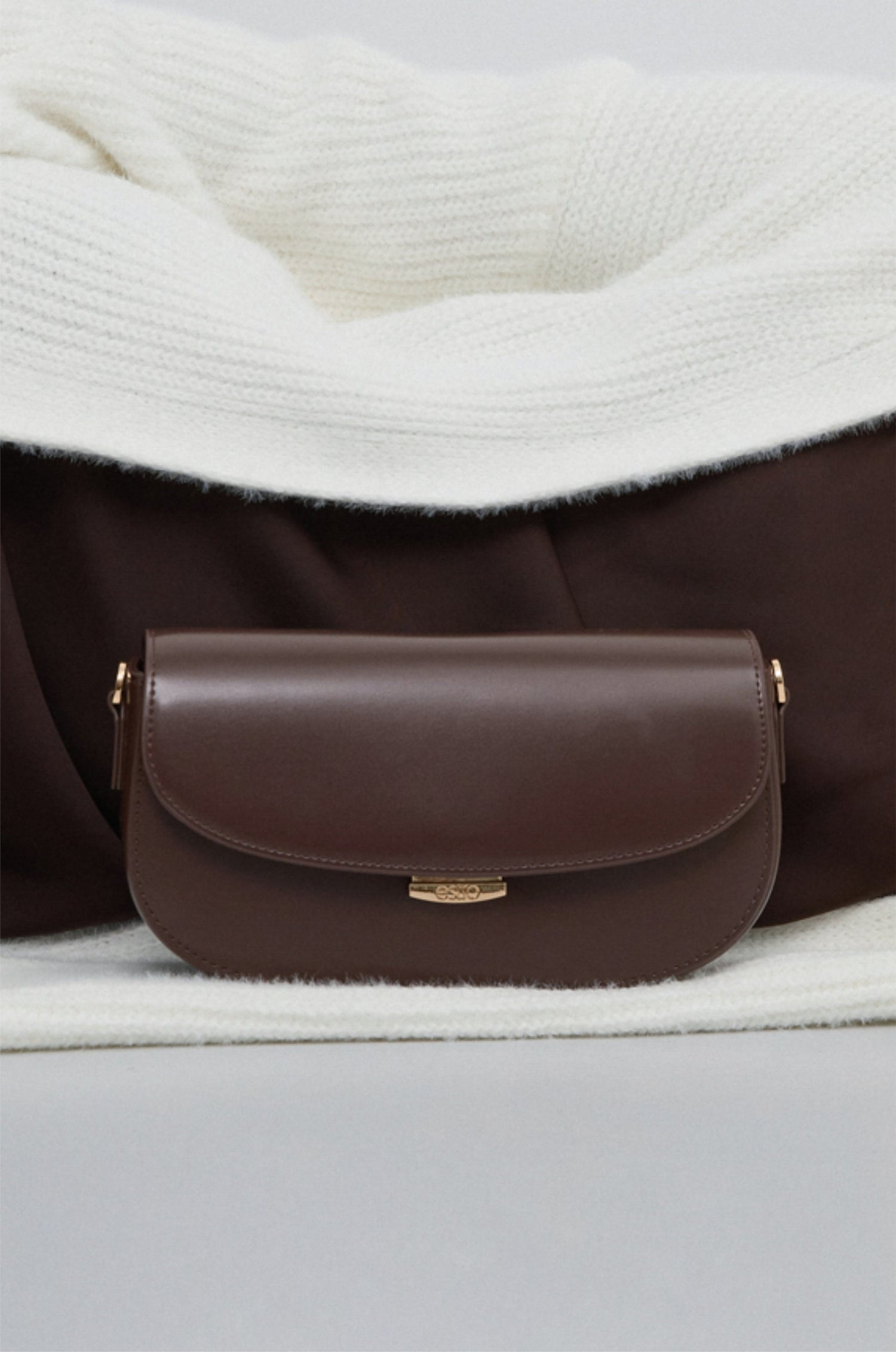 Dunkelbraune Damen-Schultertasche aus Leder in Hufeisenform auf weißem Strickstoff.