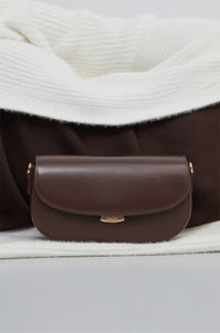 Dunkelbraune Damen-Schultertasche aus Leder in Hufeisenform auf weißem Strickstoff.