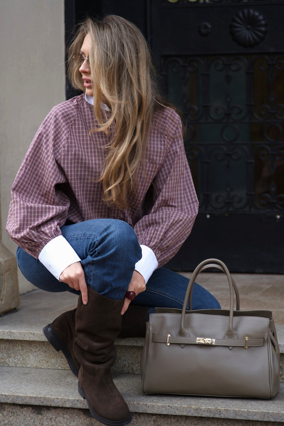 Eine Frau im Casual-Look mit karierter Bluse und Jeans hockt auf Steinstufen, während die große braune Henkeltasche neben ihr auf dem Boden steht