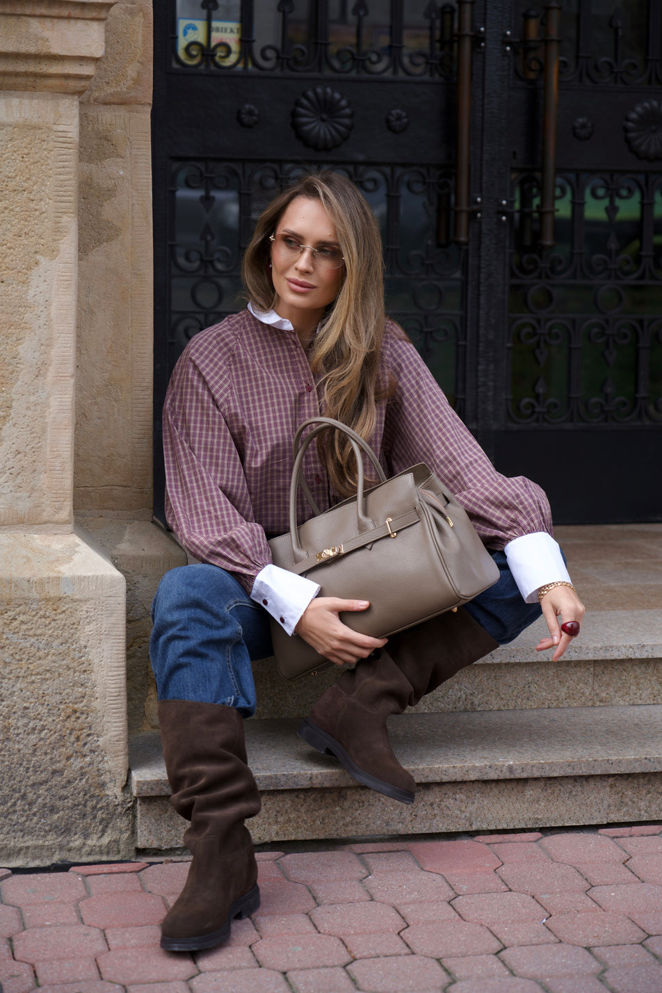 Eine Frau im herbstlichen Freizeitlook sitzt auf Steinstufen vor einem historischen Hauseingang und hält die große braune Henkeltasche aus Leder auf dem Schoß