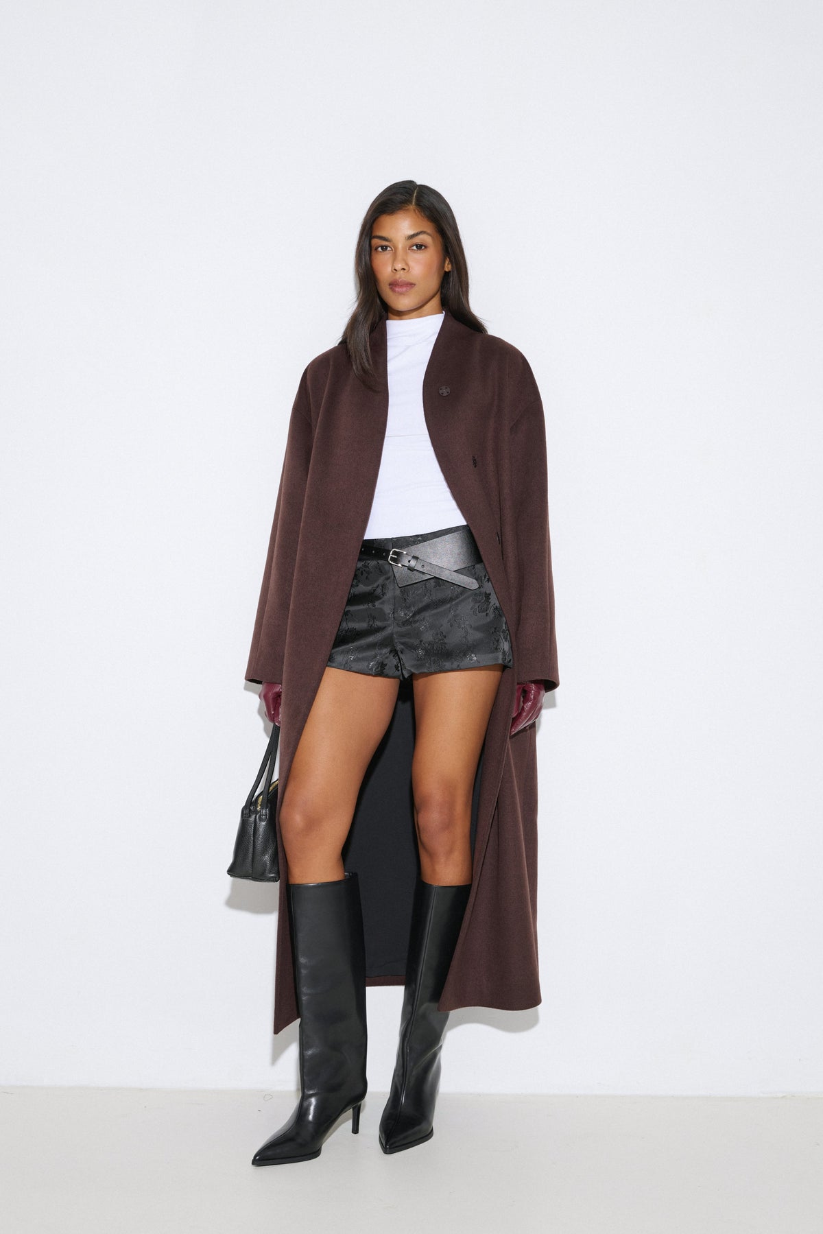 Ganzkörperansicht des offenen, langen dunkelbraunen Mantels, kombiniert mit weißem Rollkragenpullover und Shorts