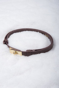 Kreative Produktinszenierung: Aufgerollter dunkelbrauner Damen-Gürtel aus Echtleder mit goldfarbenem Drehverschluss, liegend im weißen Schnee.