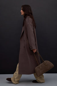 Lifestyle-Aufnahme eines Models im langen braunen Wollmantel, das die graubeige Estro-Velourstasche elegant in der Hand trägt