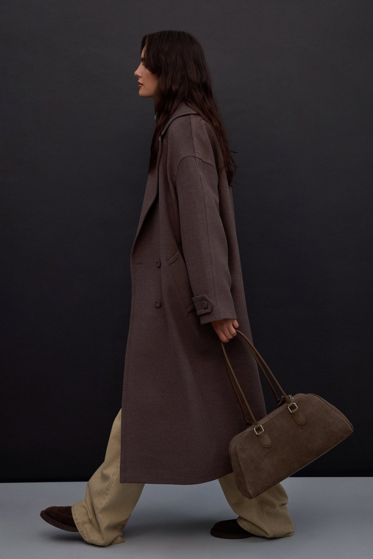 Lifestyle-Aufnahme eines Models im langen braunen Wollmantel, das die graubeige Estro-Velourstasche elegant in der Hand trägt