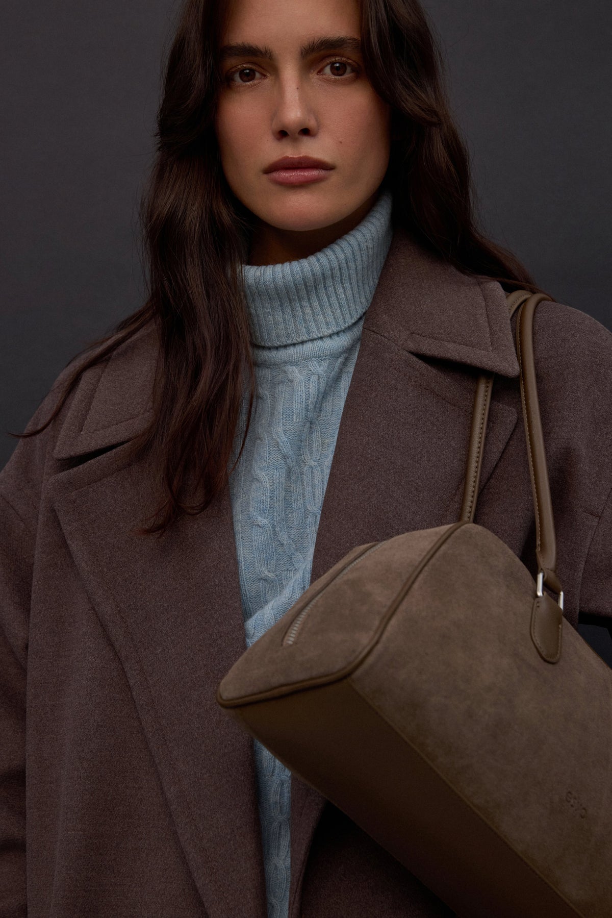 Lifestyle-Aufnahme des Models im braunen Mantel und blauen Rollkragenpullover, das die graubeige Tasche komfortabel über der Schulter trägt