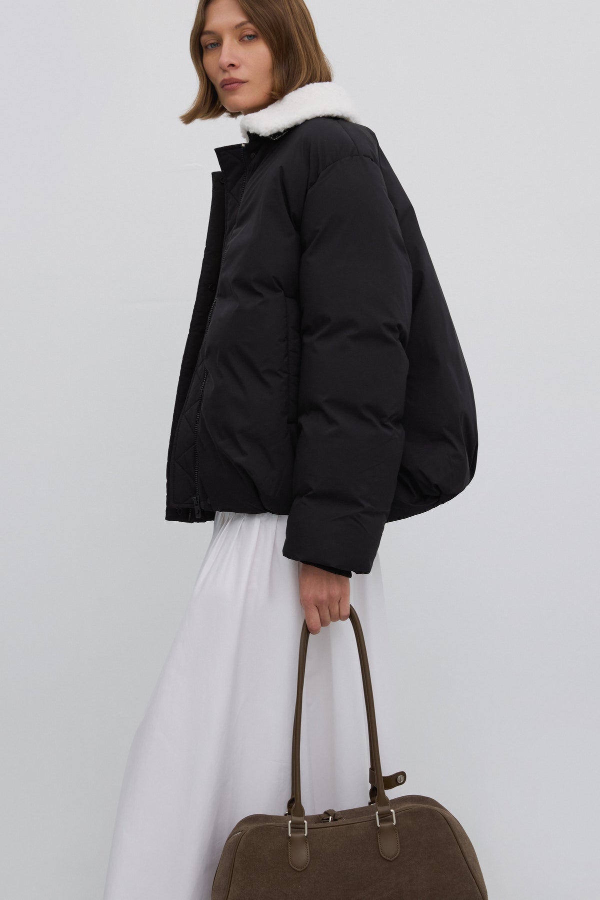 Lifestyle-Aufnahme eines Models in schwarzer Puffer-Jacke und weißem Rock, das die graubeige Velourstasche lässig in der Hand hält