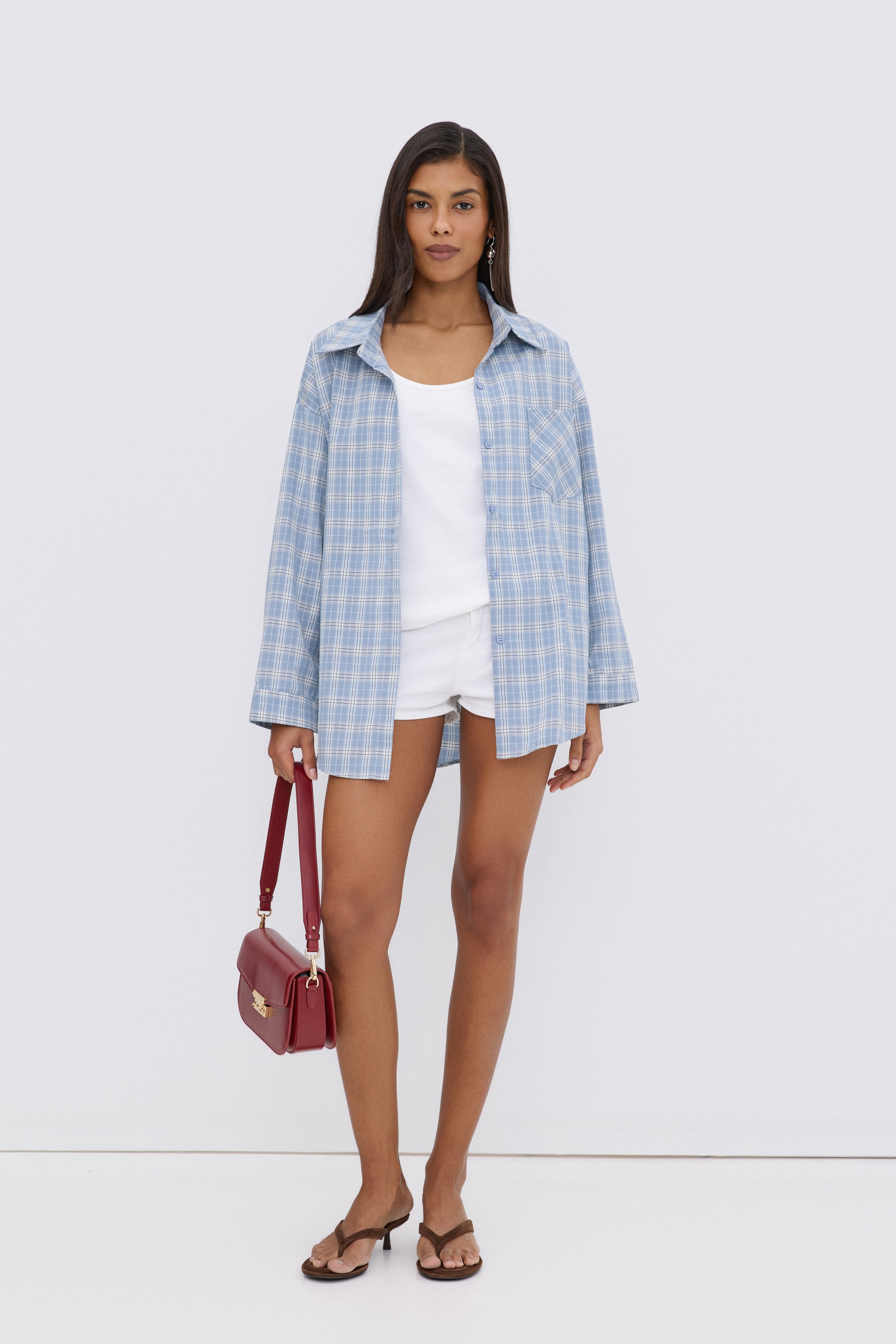 Model trägt die hellblaue Karo-Hemdbluse offen über einem weißen Top und weißen Shorts, mit einer roten Handtasche