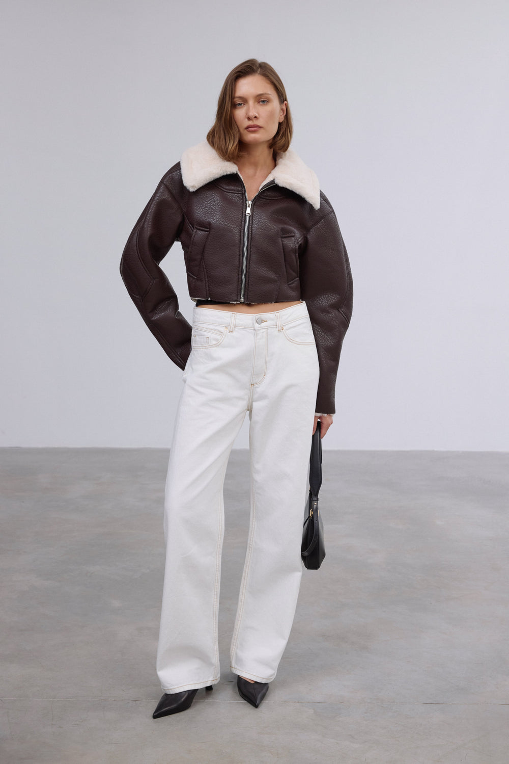 Frontalansicht des Models in der kurzen dunkelbraunen Lammfellimitat-Jacke, kombiniert mit weißer Wide-Leg-Jeans und schwarzer Handtasche