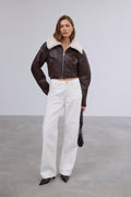 Frontalansicht des Models in der kurzen dunkelbraunen Lammfellimitat-Jacke, kombiniert mit weißer Wide-Leg-Jeans und schwarzer Handtasche