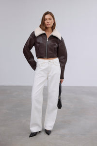 Frontalansicht des Models in der kurzen dunkelbraunen Lammfellimitat-Jacke, kombiniert mit weißer Wide-Leg-Jeans und schwarzer Handtasche