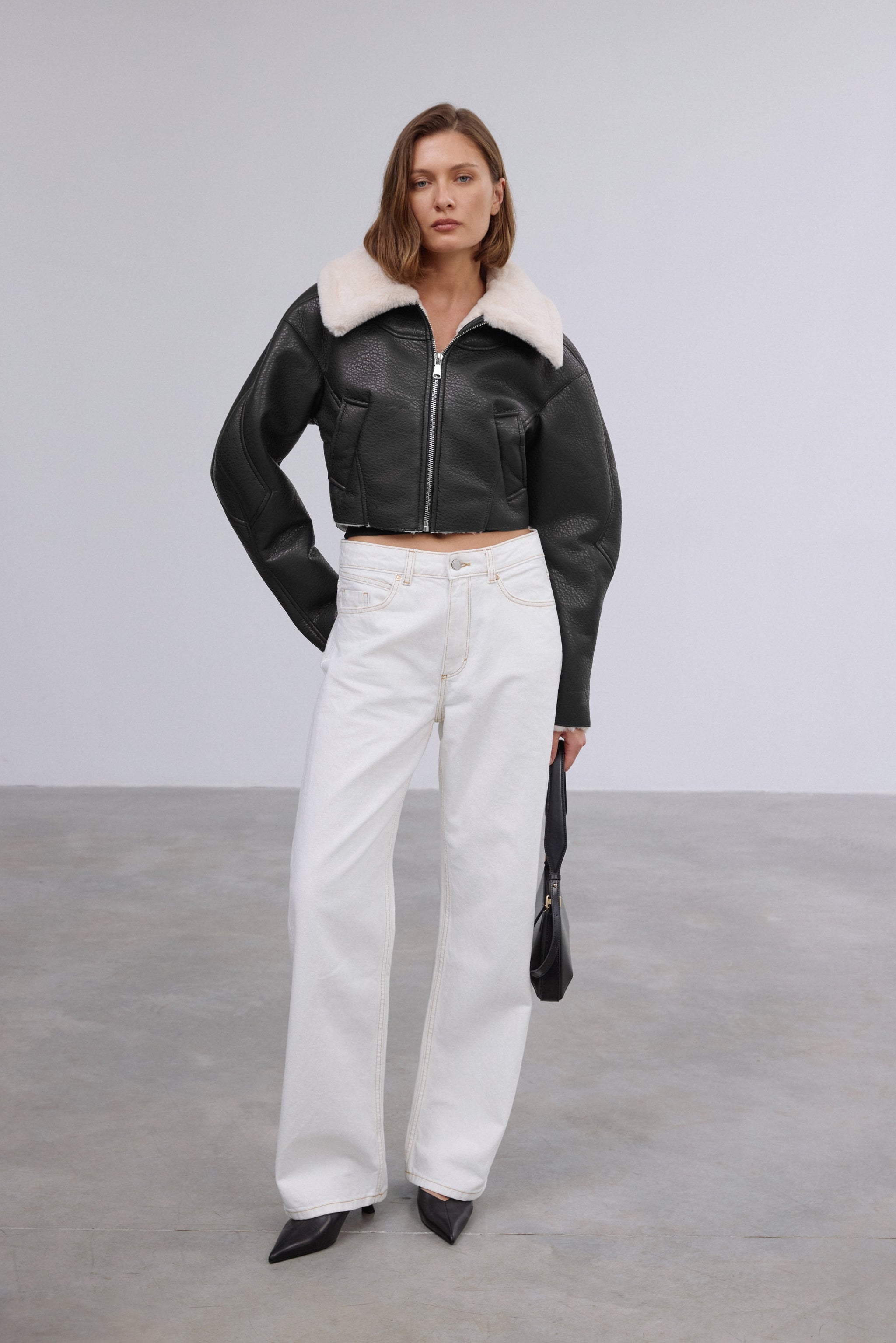 Model trägt die kurze schwarze Damenjacke aus Kunstleder in Lammfell-Optik, kombiniert mit einer weißen Hose 
