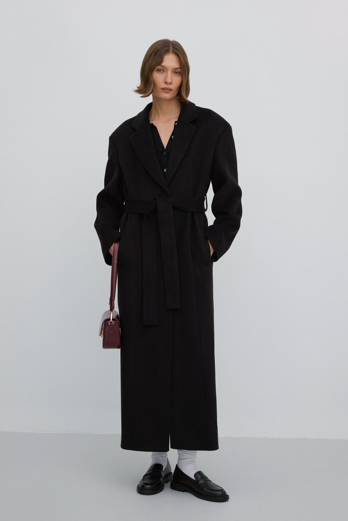 Elegante Lifestyle-Aufnahme eines Models im geschlossenen schwarzen Damen-Wollmantel mit Taillengürtel, kombiniert mit burgunderroter Handtasche und schwarzen Loafern.