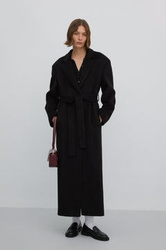 Elegante Lifestyle-Aufnahme eines Models im geschlossenen schwarzen Damen-Wollmantel mit Taillengürtel, kombiniert mit burgunderroter Handtasche und schwarzen Loafern.