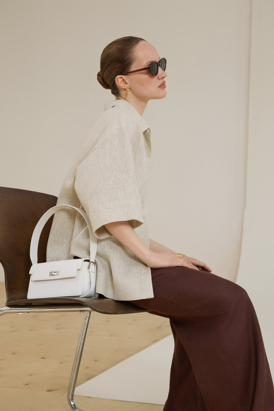 Model in einer beigen Leinenbluse sitzt auf einem Stuhl und trägt die milchweiße Schultertasche aus Echtleder