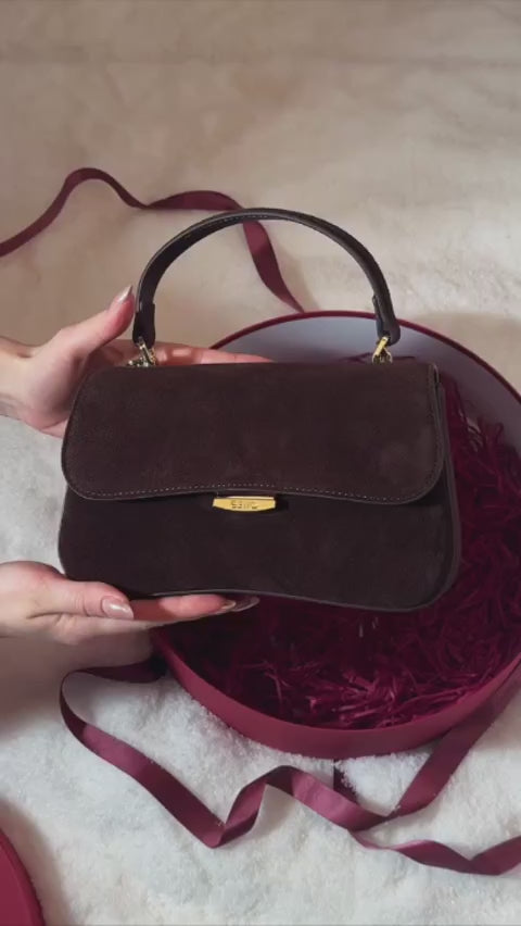 Eine Frau präsentiert die dunkelbraune Velours-Tasche elegant vor einem burgunderroten Hintergrund
