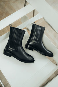 Künstlerische Ansicht des Paares schwarzer Damen-Chelsea-Boots aus Echtleder mit gerippten Stretcheinsätzen, stilvoll arrangiert auf einem weißen Stuhl   