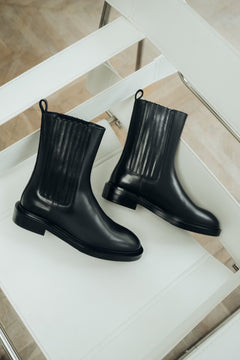 Künstlerische Ansicht des Paares schwarzer Damen-Chelsea-Boots aus Echtleder mit gerippten Stretcheinsätzen, stilvoll arrangiert auf einem weißen Stuhl   