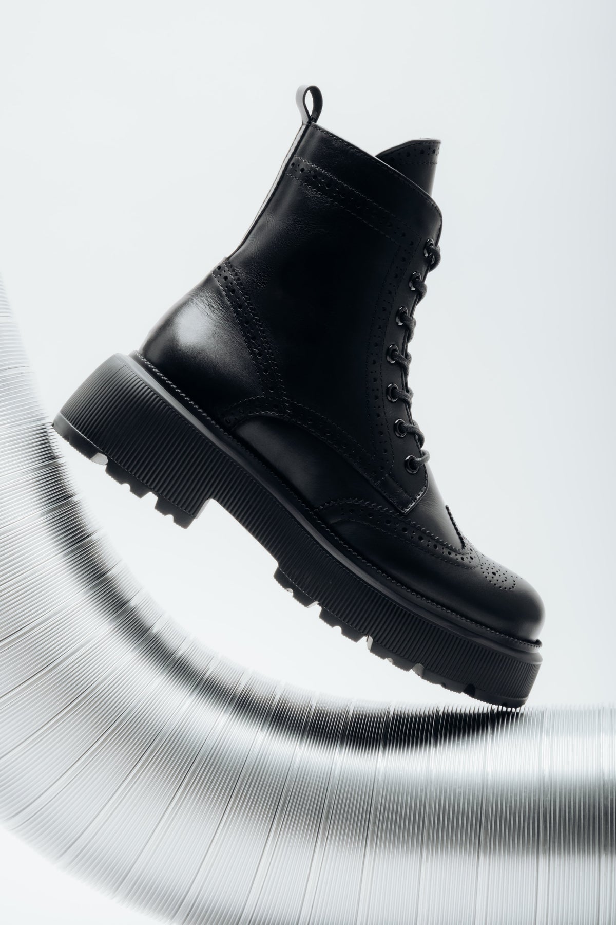 Künstlerische Seitenansicht der rechten schwarzen Damen-Schnürstiefelette aus Leder mit Brogue-Lochmuster, die dynamisch auf einem geriffelten silbernen Metallrohr präsentiert wird.   