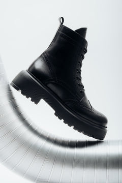 Künstlerische Seitenansicht der rechten schwarzen Damen-Schnürstiefelette aus Leder mit Brogue-Lochmuster, die dynamisch auf einem geriffelten silbernen Metallrohr präsentiert wird.   
