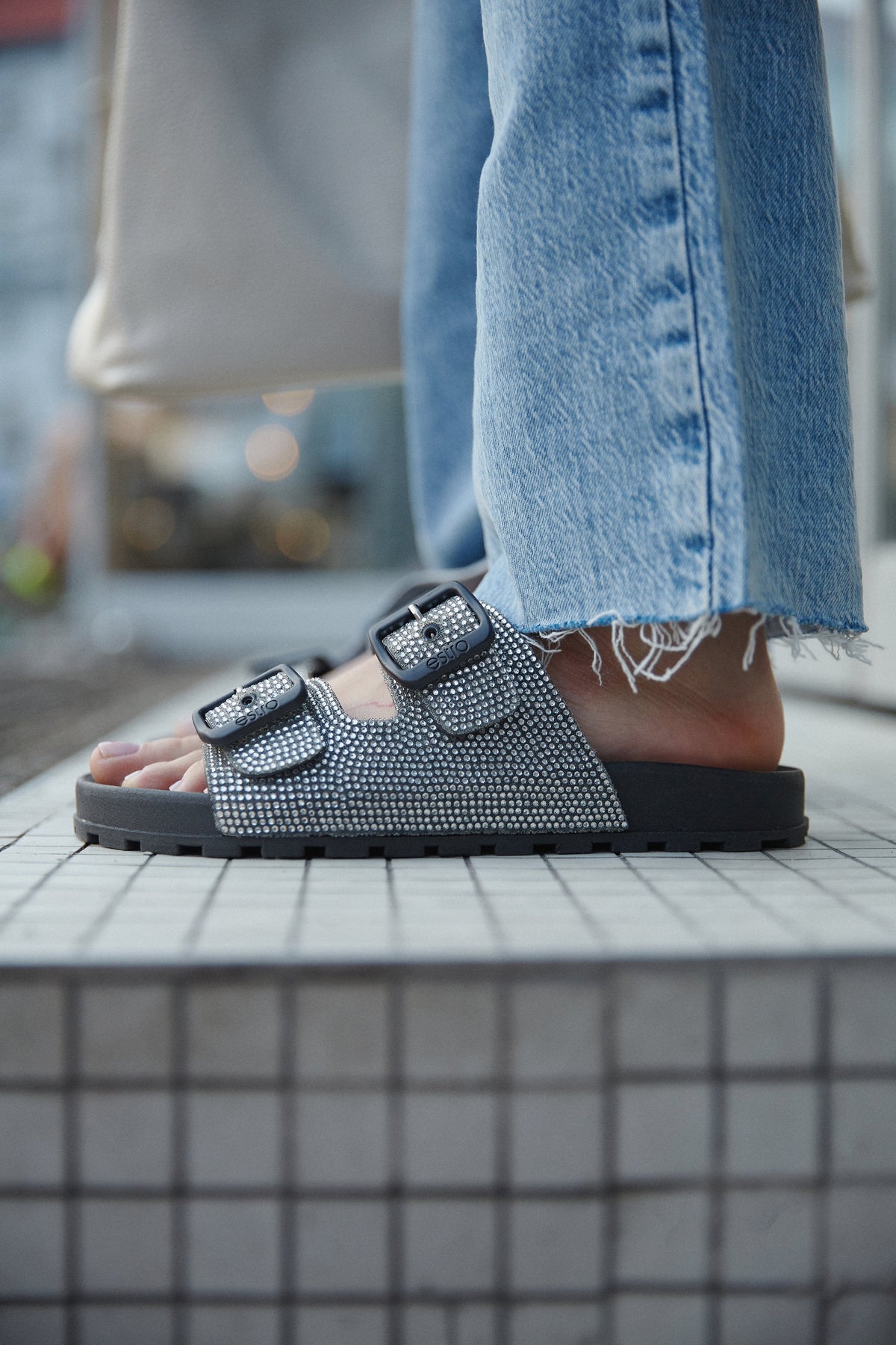 Lifestyle-Aufnahme eines Models in hellen Jeans, das die schwarzen Pantoletten mit funkelnden Zirkonia-Riemen trägt