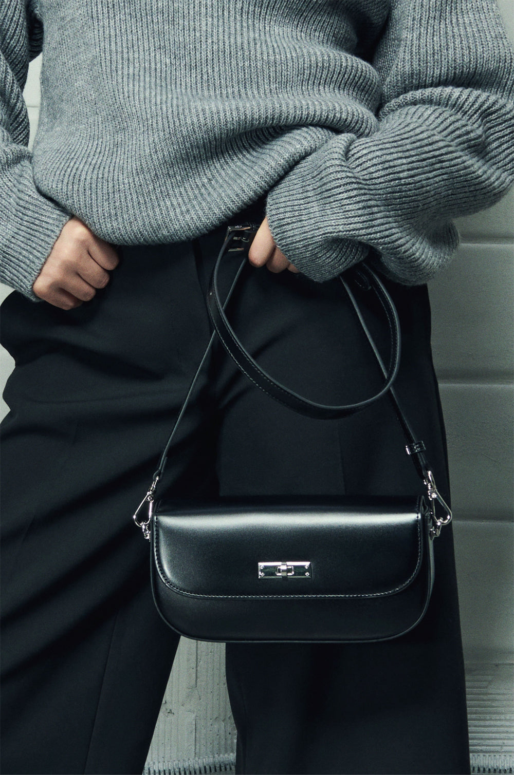 Model im grauen Pullover hält die schwarze Leder-Schultertasche mit dem langen Lederriemen