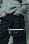 Model im grauen Pullover hält die schwarze Leder-Schultertasche mit dem langen Lederriemen