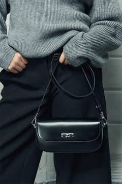 Model im grauen Pullover hält die schwarze Leder-Schultertasche mit dem langen Lederriemen