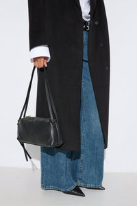 Stilvolle Lifestyle-Aufnahme eines Models im langen schwarzen Mantel, weiten Jeans und mit der schwarzen Schultertasche aus italienischem Echtleder.