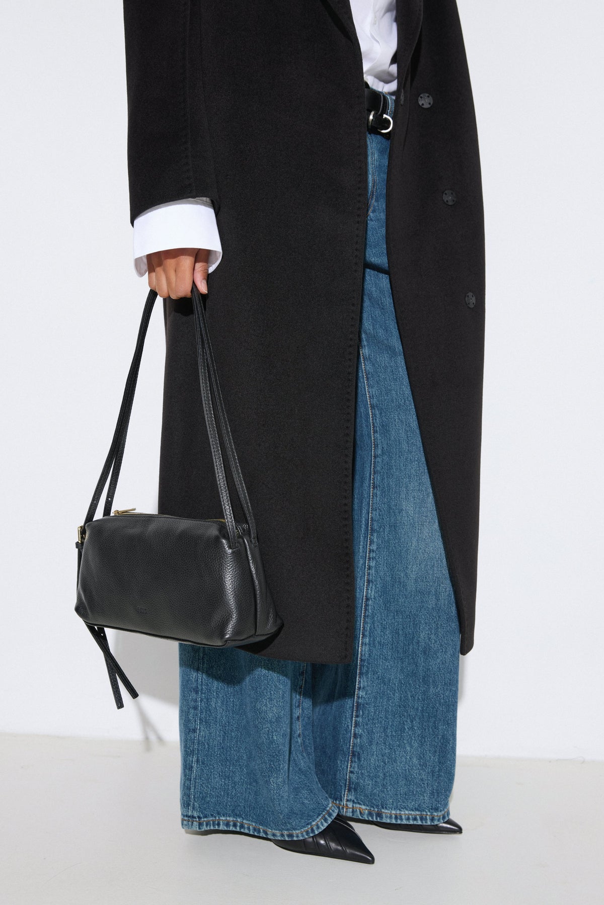 Stilvolle Lifestyle-Aufnahme eines Models im langen schwarzen Mantel, weiten Jeans und mit der schwarzen Schultertasche aus italienischem Echtleder.