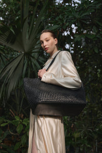 Ein Model in einer üppigen grünen Kulisse trägt die große schwarze Shopper-Tasche aus geflochtenem Leder.