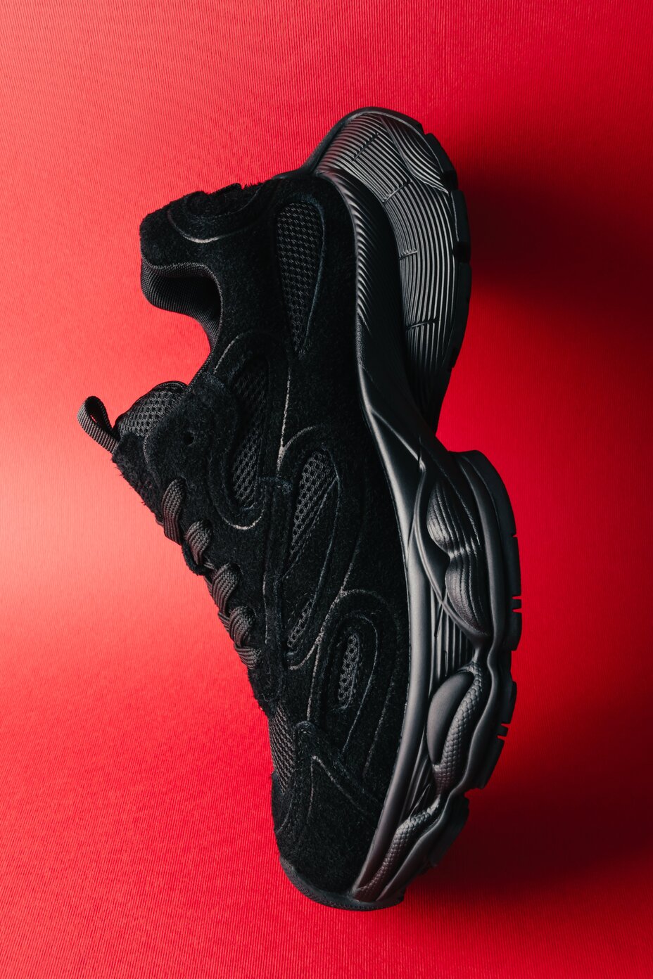 Seitliche Ansicht des schwarzen Chunky-Sneakers aus Veloursleder mit Mesh-Einsätzen vor rotem Hintergrund