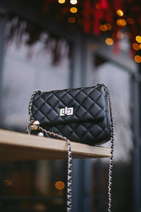 Künstlerische Lifestyle-Aufnahme der schwarzen Damen-Stepptasche mit silberner Kette vor abendlicher Stadtkulisse