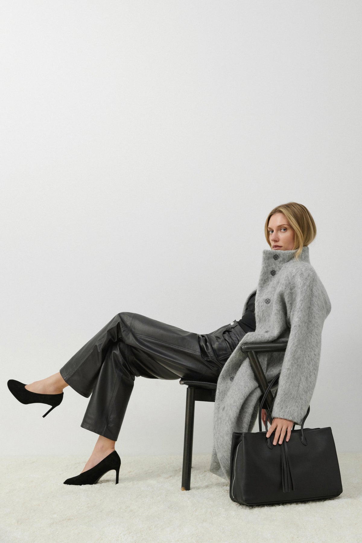 Model im grauen Mantel sitzt auf einem Stuhl neben der geräumigen schwarzen Leder-Tote-Bag