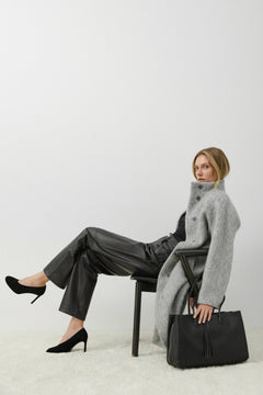 Model im grauen Mantel sitzt auf einem Stuhl neben der geräumigen schwarzen Leder-Tote-Bag