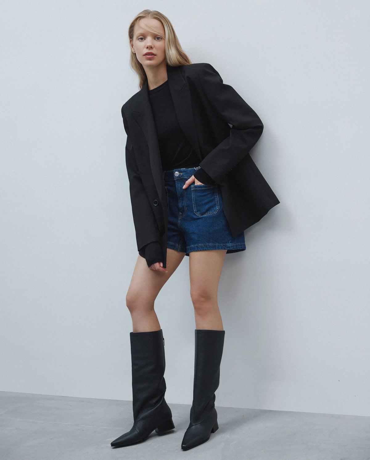 Model im schwarzen Blazer und Denim-Shorts präsentiert schwarze Lederstiefel mit weitem Schaft
