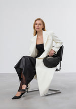 Model im weißen Mantel und schwarzen Kleid sitzt auf einem Stuhl und hält die schwarze Halbmond-Tasche elegant in der Hand