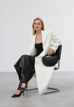 Model im weißen Mantel und schwarzen Kleid sitzt auf einem Stuhl und hält die schwarze Halbmond-Tasche elegant in der Hand